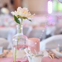 décoration de table avec ancienne bouteille de lait remplie d'eau et de billes rose, d'un pot de confiture avec sable gris servant de porte bougie et d'une grosse bougie rose décorée avec de la toile de jute et de la dentelle