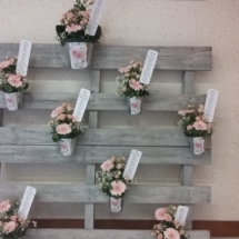 plan de table fabriqué dans une palette peinte en gris et légèrement patinée blanc avec vissés dessus des petits pots de fleurs en zinc avec le numéro des tables et le nom des invités