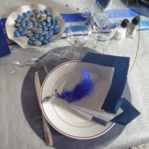 pliage de deux serviettes en forme d'éventail, une de couleur bleue et l'autre de couleur argenté, le tout tenu par une plume bleue