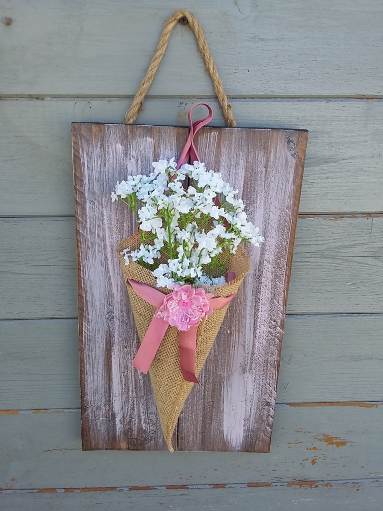 cône de fleurs en toile de jute avec une fleur en tissus posé sur sur un noeud en satin rose et le tout fixé sur un cadre en bois teinté et légèrement patiné rose tendre.