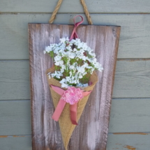 cône de fleurs en toile de jute avec une fleur en tissus posé sur sur un noeud en satin rose et le tout fixé sur un cadre en bois teinté et légèrement patiné rose tendre.