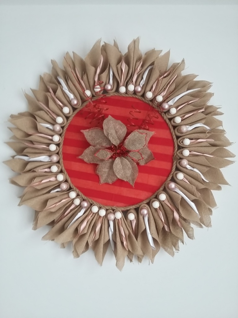 grande couronne de noël en toile de jute en forme de soleil avec au milieu de la toile de jute couleur rouge, une fleur en toile de jute naturel et fils torsadés rouge pailletés. La couronne fait 80 cm de diamètre