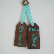 plaque de porte en bois teinté chêne foncé avec inscriptions bienvenue chez nous de couleur bleu turquoise, le tout tenu par un ruban de tissu vichy turquoise et une fleur en toile de jute fait main