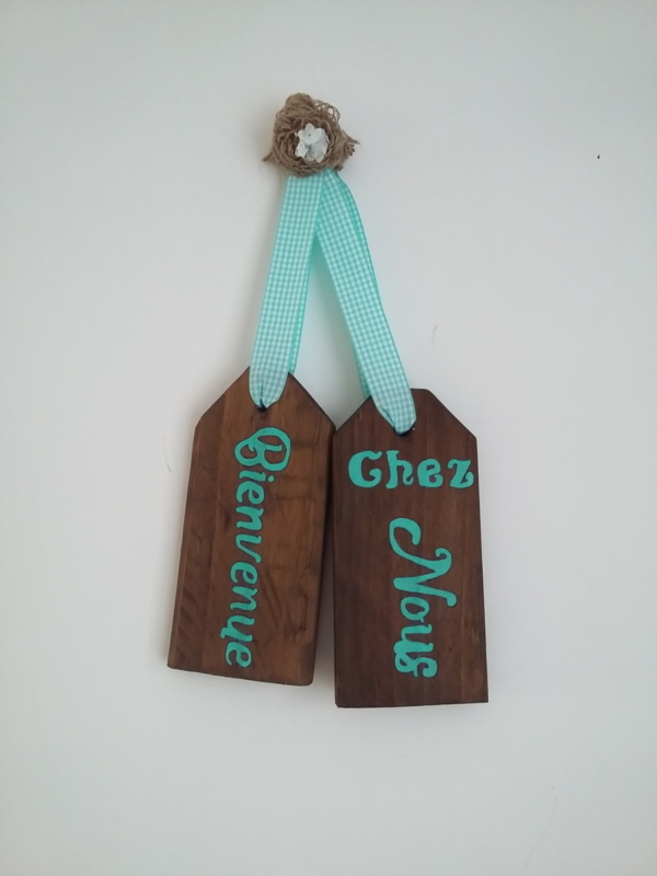 plaque de porte en bois teinté chêne foncé avec inscriptions bienvenue chez nous de couleur bleu turquoise, le tout tenu par un ruban de tissu vichy turquoise et une fleur en toile de jute fait main