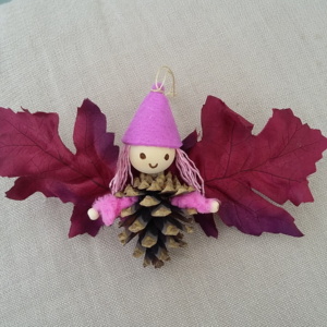 petit lutin ailé à suspendre fabriqué avec des pommes de pin, une perle de bois pour la tête, des feuilles de couleur rouge vin pour les ailes et un petit chapeau pointu de couleur mauve.