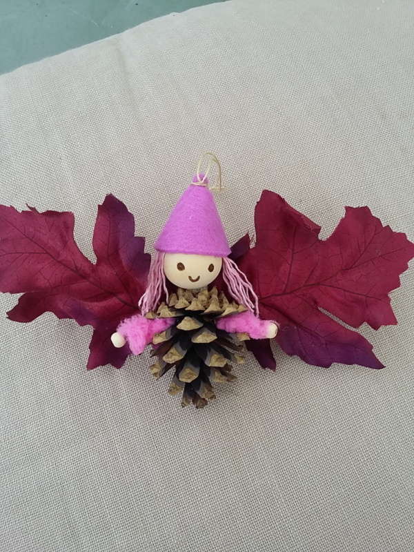 petit lutin ailé à suspendre fabriqué avec des pommes de pin, une perle de bois pour la tête, des feuilles de couleur rouge vin pour les ailes et un petit chapeau pointu de couleur mauve.