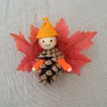 petit lutin ailé à suspendre fabriqué avec des pommes de pin, une perle de bois pour la tête, des feuilles de couleur rouge pour les ailes et un petit chapeau pointu de couleur orange.