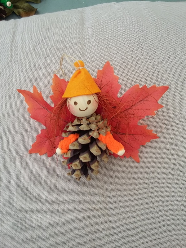 petit lutin ailé à suspendre fabriqué avec des pommes de pin, une perle de bois pour la tête, des feuilles de couleur rouge pour les ailes et un petit chapeau pointu de couleur orange.