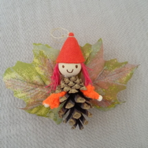 petit lutin ailé à suspendre fabriqué avec des pommes de pin, une perle de bois pour la tête, des feuilles de couleur mauve servant d'ailes et un petit chapeau pointu de couleur rose pâle.