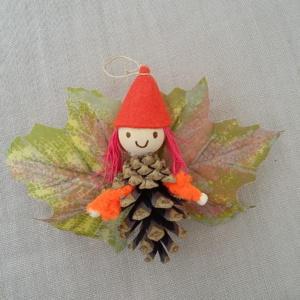 petit lutin ailé à suspendre fabriqué avec des pommes de pin, une perle de bois pour la tête, des feuilles de couleur mauve servant d'ailes et un petit chapeau pointu de couleur rose pâle.