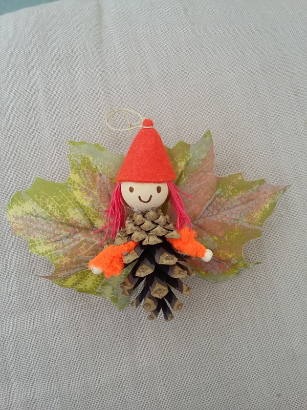 petit lutin ailé à suspendre fabriqué avec des pommes de pin, une perle de bois pour la tête, des feuilles de couleur mauve servant d'ailes et un petit chapeau pointu de couleur rose pâle.