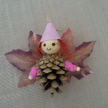 petit lutin ailé à suspendre fabriqué avec des pommes de pin, une perle de bois pour la tête, des feuilles de couleur mauve servant d'ailes et un petit chapeau pointu de couleur rose pâle.