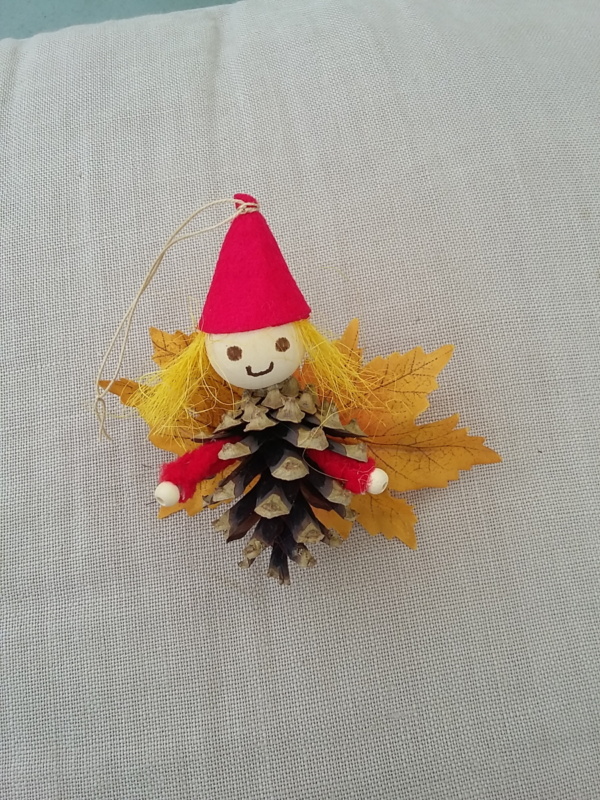 petit lutin ailé à suspendre fabriqué avec des pommes de pin, une perle de bois pour la tête, des feuilles de couleur jaune pour les ailes et un petit chapeau pointu de couleur rouge