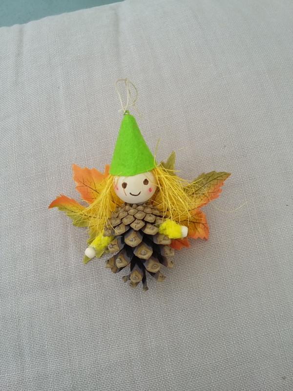 petit lutin ailé à suspendre fabriqué avec des pommes de pin, une perle de bois pour la tête, des feuilles de couleur orange jaune et vert pour les ailes et un petit chapeau pointu de couleur vert