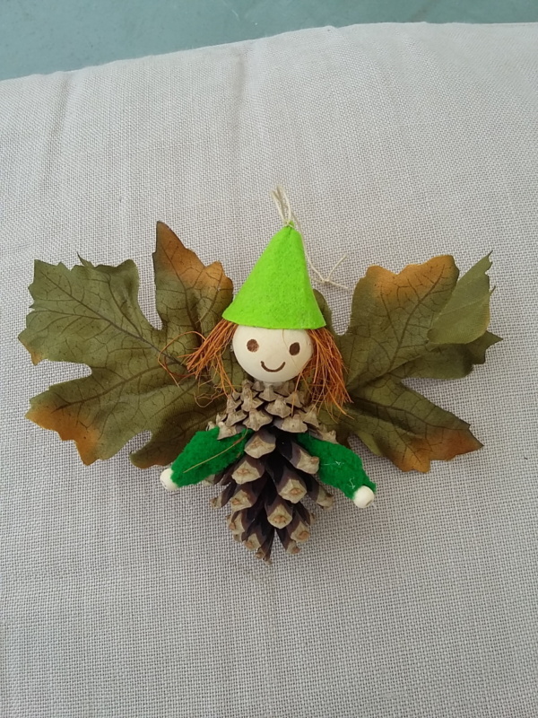 petit lutin ailé à suspendre fabriqué avec des pommes de pin, une perle de bois pour la tête, des feuilles de couleur vert foncé pour les ailes et un petit chapeau pointu de couleur vert clair