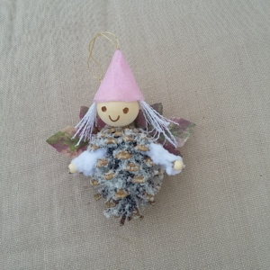 petit lutin ailé à suspendre fabriqué avec une pomme de pin enneigé, une perle de bois pour la tête, des feuilles de couleur violet et vert pour les ailes et un petit chapeau pointu de couleur rose pâle