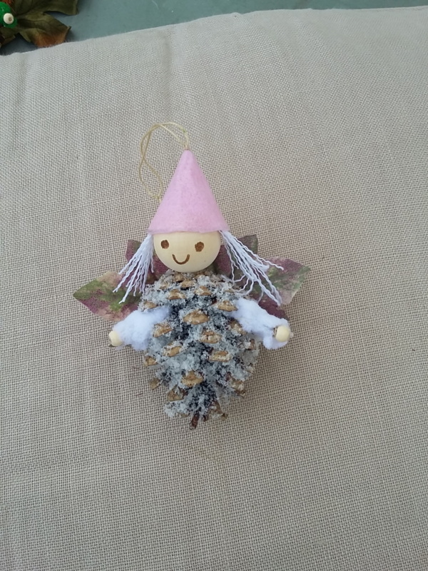 petit lutin ailé à suspendre fabriqué avec une pomme de pin enneigé, une perle de bois pour la tête, des feuilles de couleur violet et vert pour les ailes et un petit chapeau pointu de couleur rose pâle