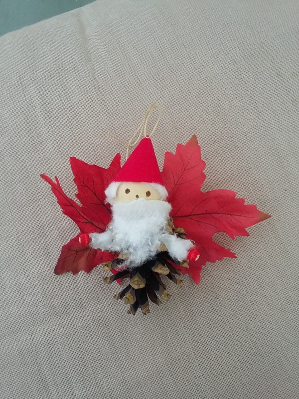 petit lutin père noël ailé à suspendre fabriqué avec des pommes de pin, une perle de bois pour la tête, des feuilles de couleur rouge pour les ailes et un petit chapeau pointu de couleur rouge.