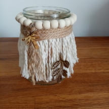 Petit bougeoir en macramé avec autour des perles de bois et un ruban de toile de jute . Une petite libellule en bois est collée par dessus et un ornement en métal est suspendu sur le côté.