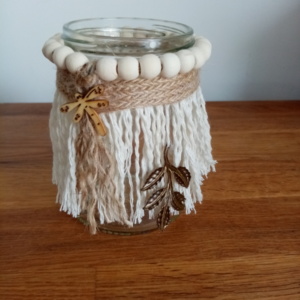 Petit bougeoir en macramé avec autour des perles de bois et un ruban de toile de jute . Une petite libellule en bois est collée par dessus et un ornement en métal est suspendu sur le côté.