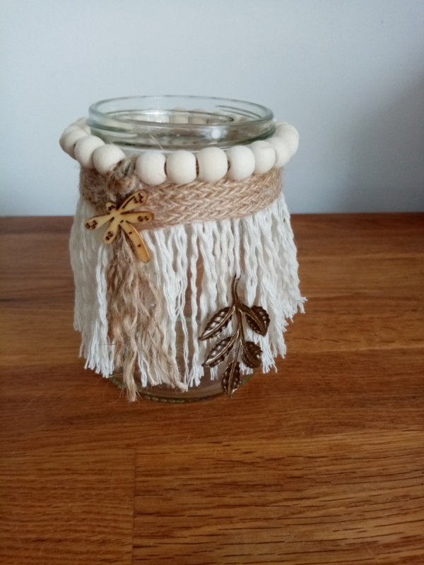 Petit bougeoir en macramé avec autour des perles de bois et un ruban de toile de jute . Une petite libellule en bois est collée par dessus et un ornement en métal est suspendu sur le côté.