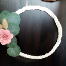 Couronne de macramé tressé avec fleur en bois de couleur rose et des petites feuilles d'eucalyptus.