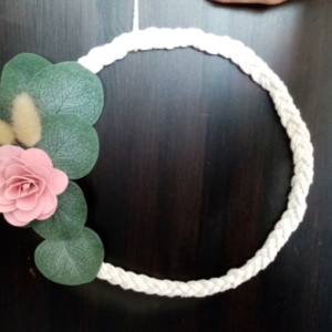 Couronne de macramé tressé avec fleur en bois de couleur rose et des petites feuilles d'eucalyptus.