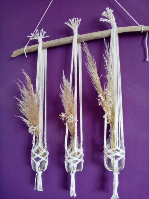 Trois soliflores en macramé suspendus sur une branche de bois flotté. Des fleurs de pampa et du gypsophile séché agrémentent ces soliflores