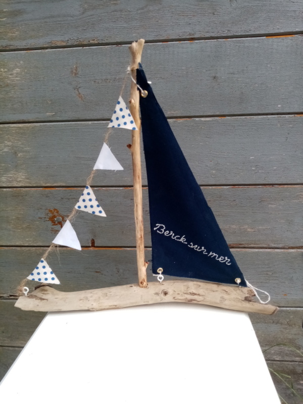 Bateau en bois flotté avec une voile bleu marine et une inscription berck sur mer en broderie fait main ainsi qu'une guirlande de petits fanions tissus blanc et petits pois bleus