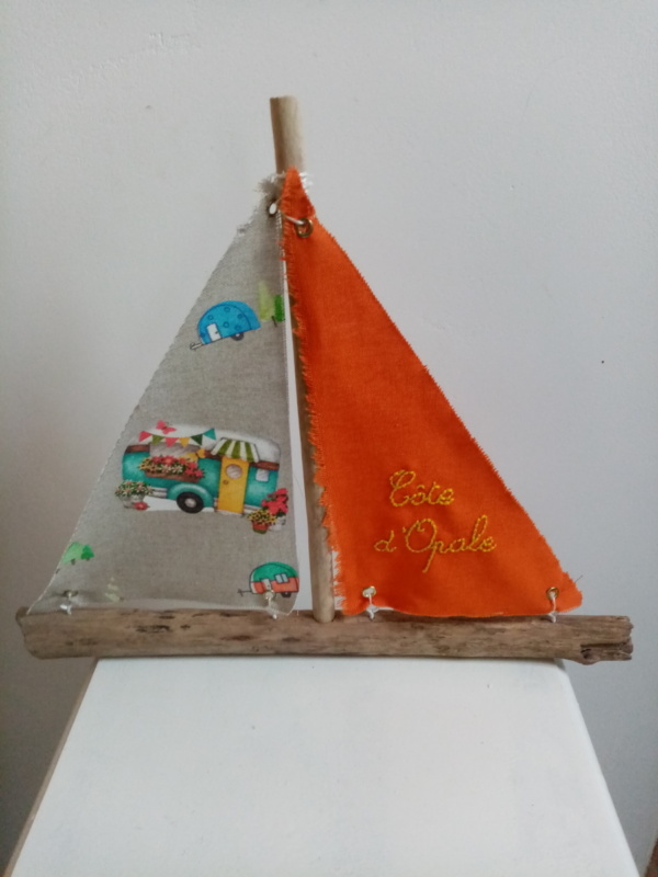 Bateau en bois flotté avec deux voiles. Une voile couleur orange et une inscription " Côte d'Opale" en broderie jaune et une autre voile avec des imprimés combi.