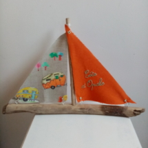 Bateau en bois flotté avec deux voiles. Une voile couleur orange et une inscription " Côte d'Opale" en broderie bleu clair et une autre voile avec des imprimés de caravanes vintage.