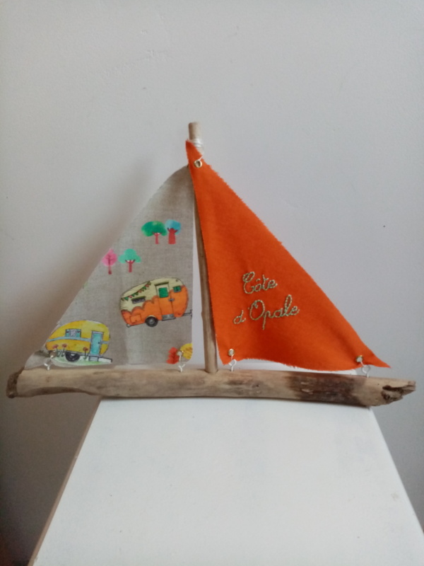 Bateau en bois flotté avec deux voiles. Une voile couleur orange et une inscription " Côte d'Opale" en broderie bleu clair et une autre voile avec des imprimés de caravanes vintage.