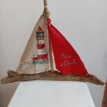 Bateau en bois flotté avec deux voiles. Une voile rouge et une inscription " Côte d'Opale" en broderie bleu clair et une autre voile avec un imprime d'un phare.